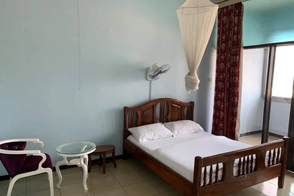A bedroom with a wooden bed, white bedding, a small round table, a chair, a standing fan, and a window with curtains.