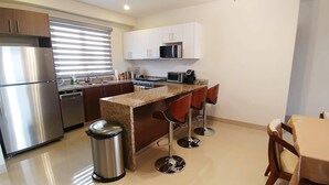 Une cuisine moderne équipée d’un réfrigérateur en acier inoxydable, d’un four à micro-ondes, d’une cuisinière et d’un évier. On y trouve deux tabourets de bar et une table recouverte d’une nappe beige.
