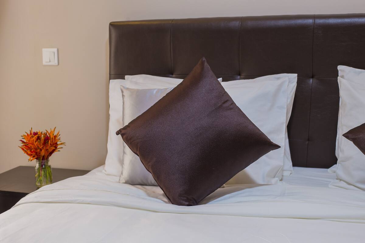 A bed with white bedding and a dark brown headboard, a vase with flowers on a nightstand, and a plain wall.