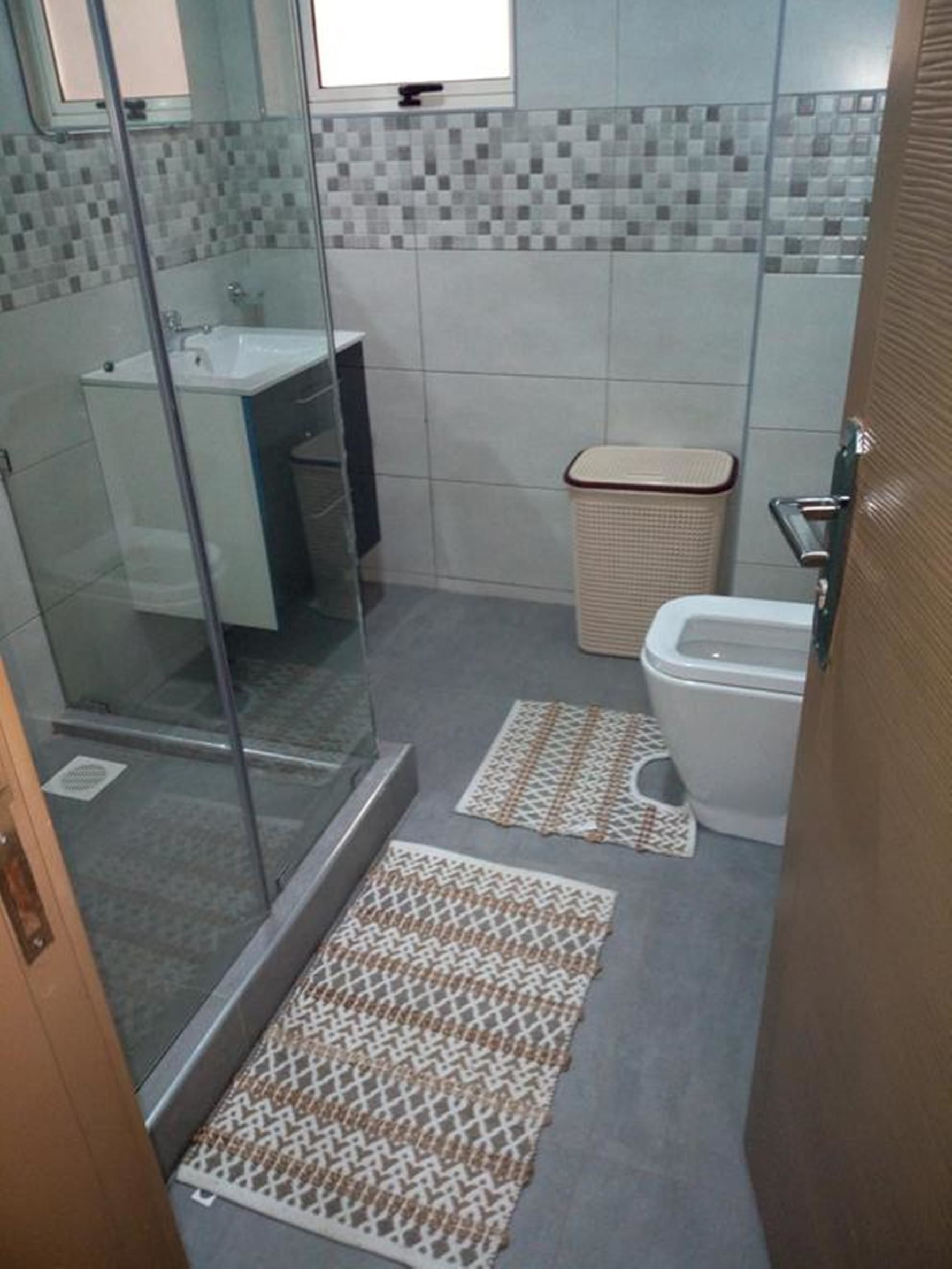 A bathroom with a glass shower enclosure, a white sink, a toilet, and patterned mats on the floor.