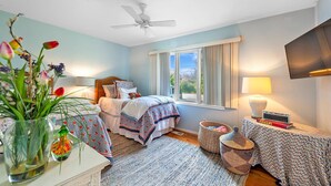 Ein Schlafzimmer mit einem Bett, einem Deckenventilator, einem Fenster mit Vorhängen, einem Nachttisch mit Lampe und einem an der Wand montierten Fernseher.