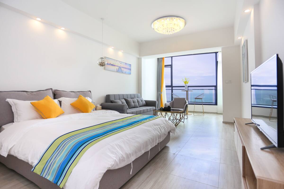 A modern hotel room with a large bed, a gray sofa, a small table, and a TV.