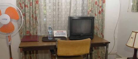 A room with a desk, a television, a fan, a chair, and a lamp.