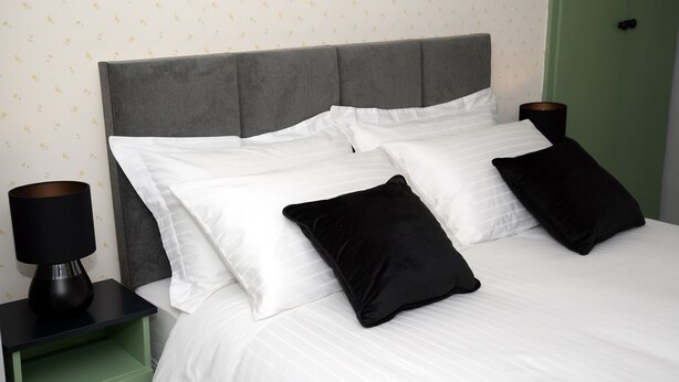 A neatly made bed with white linens and black pillows, flanked by two black nightstands with lamps, against a wall with a subtle pattern.