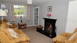 A cozy living room with a fireplace, a sofa set, a small table with a bottle, and a window with curtains.