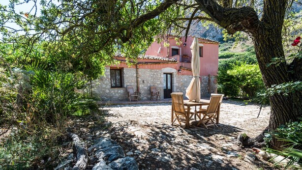 A stone building with a tiled roof, a patio with a table and chairs, and a large tree in the foreground. - Marotta Chalet, on the slopes of Erice (Valderice)