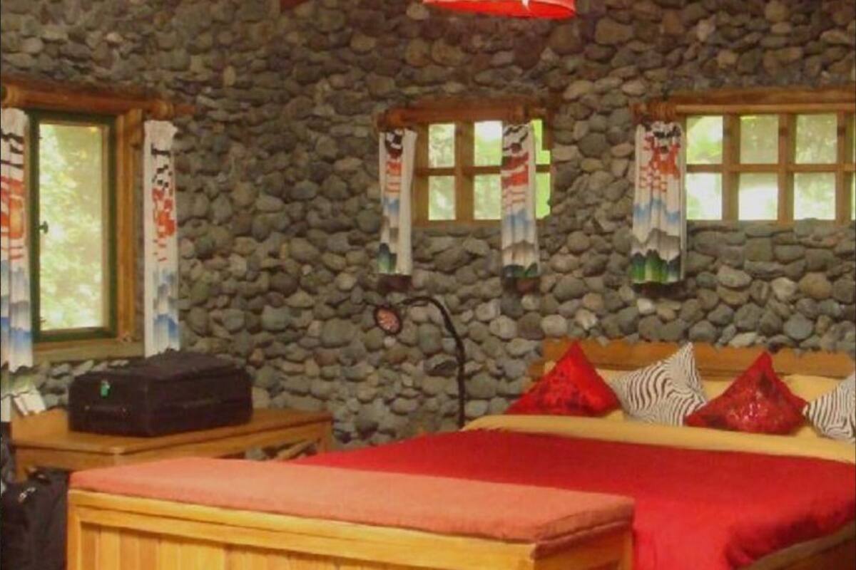 Um quarto com parede de pedra, cama com roupa de cama vermelha, um banco de madeira e uma mala sobre uma mesa.