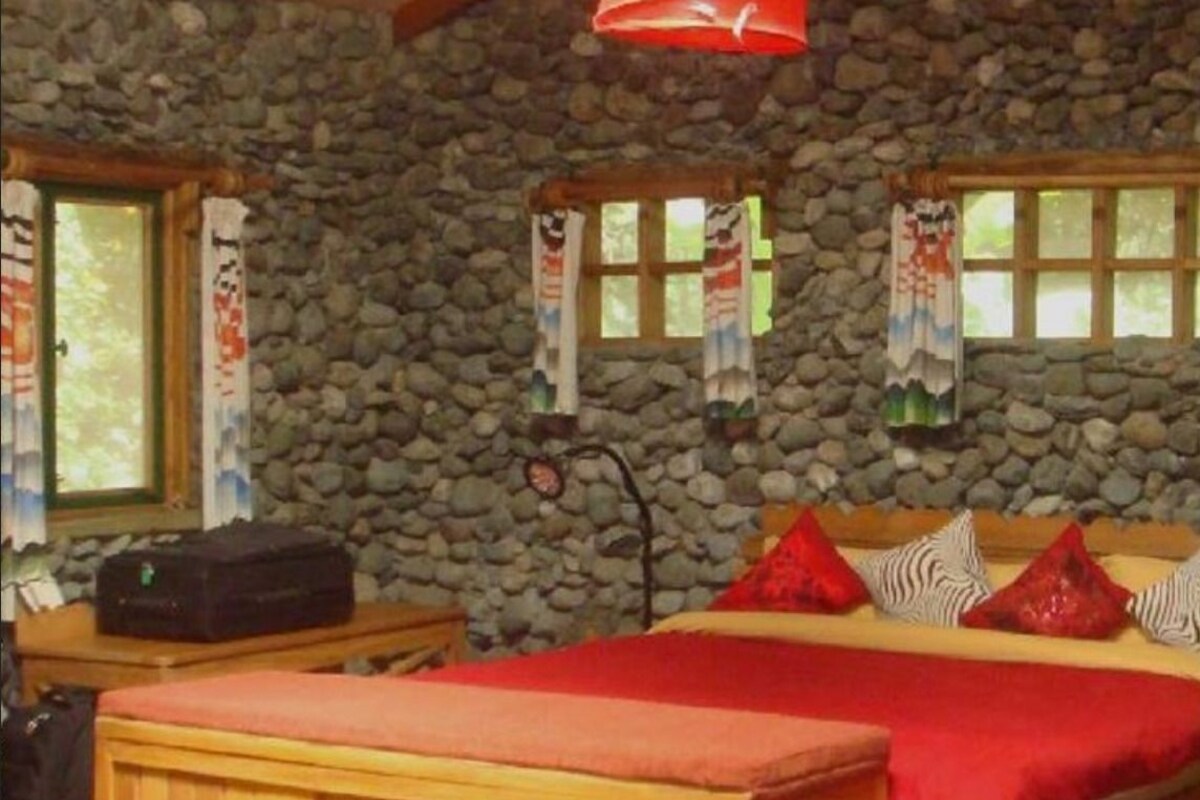 Una habitación con pared de piedra, cama con ropa de cama roja, un banco de madera y una maleta sobre una mesa.