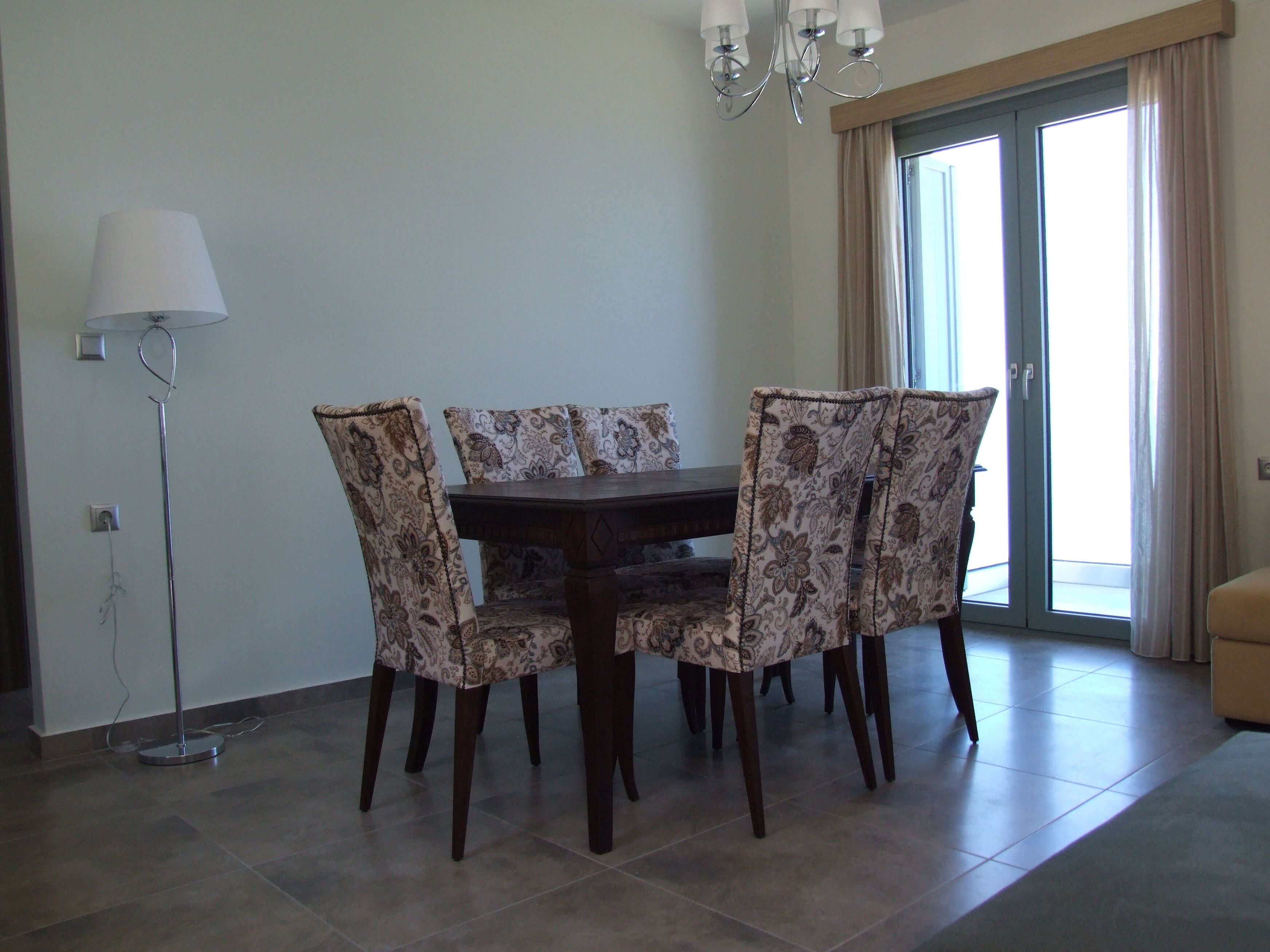 A dining area with a table and chairs, a floor lamp, and a view of the outside through a window.