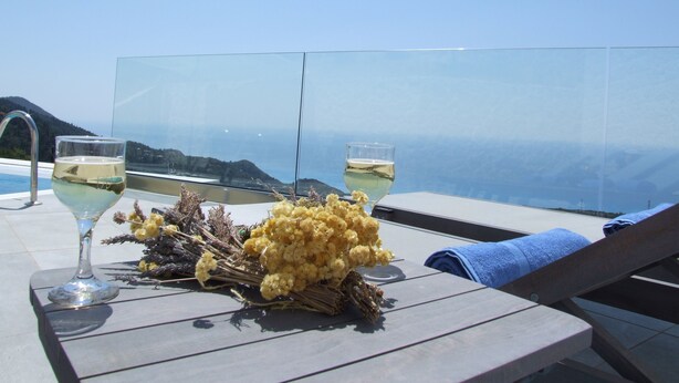 Ein Tisch mit zwei Weingläsern, einem Strauß gelber Blumen und einem blauen Handtuch auf einem Balkon mit Blick auf ein Gewässer und Hügel.