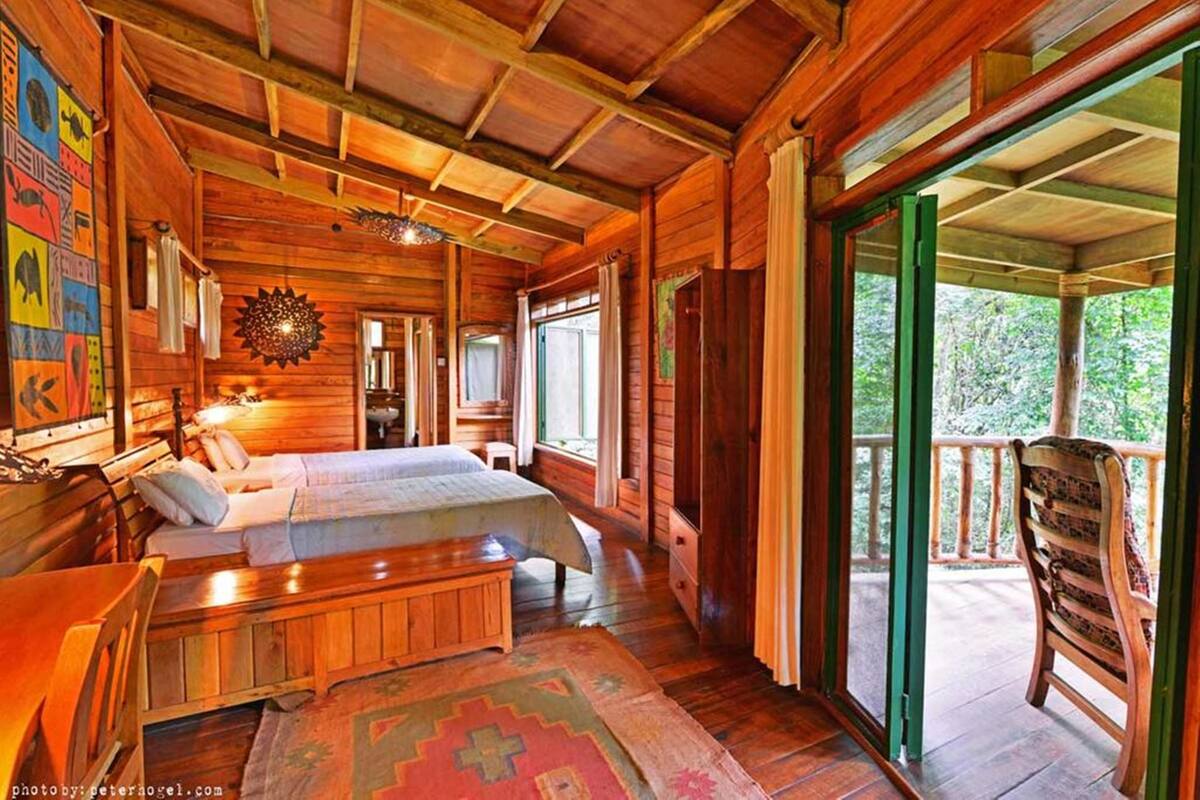 Uma cabana de madeira com uma cama grande, um banco e uma varanda com vista para vegetação.