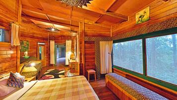Interior acogedor de una cabaña con una cama grande, un banco y una ventana con vistas al bosque.