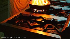 A chef in a white uniform with a pot on a stove emitting flames. - The Rainforest Geo Lodge (Lugazi)