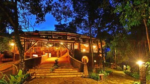 A well-lit outdoor area with a staircase leading to a covered structure, surrounded by trees and plants. - The Rainforest Geo Lodge (Lugazi)