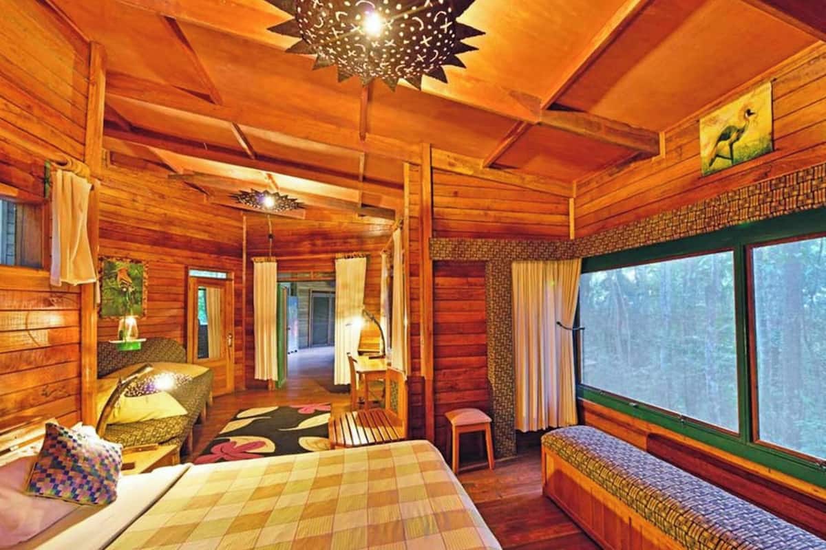 Interior de uma cabana de tronco com uma janela grande, uma cama, um banco e um lustre.