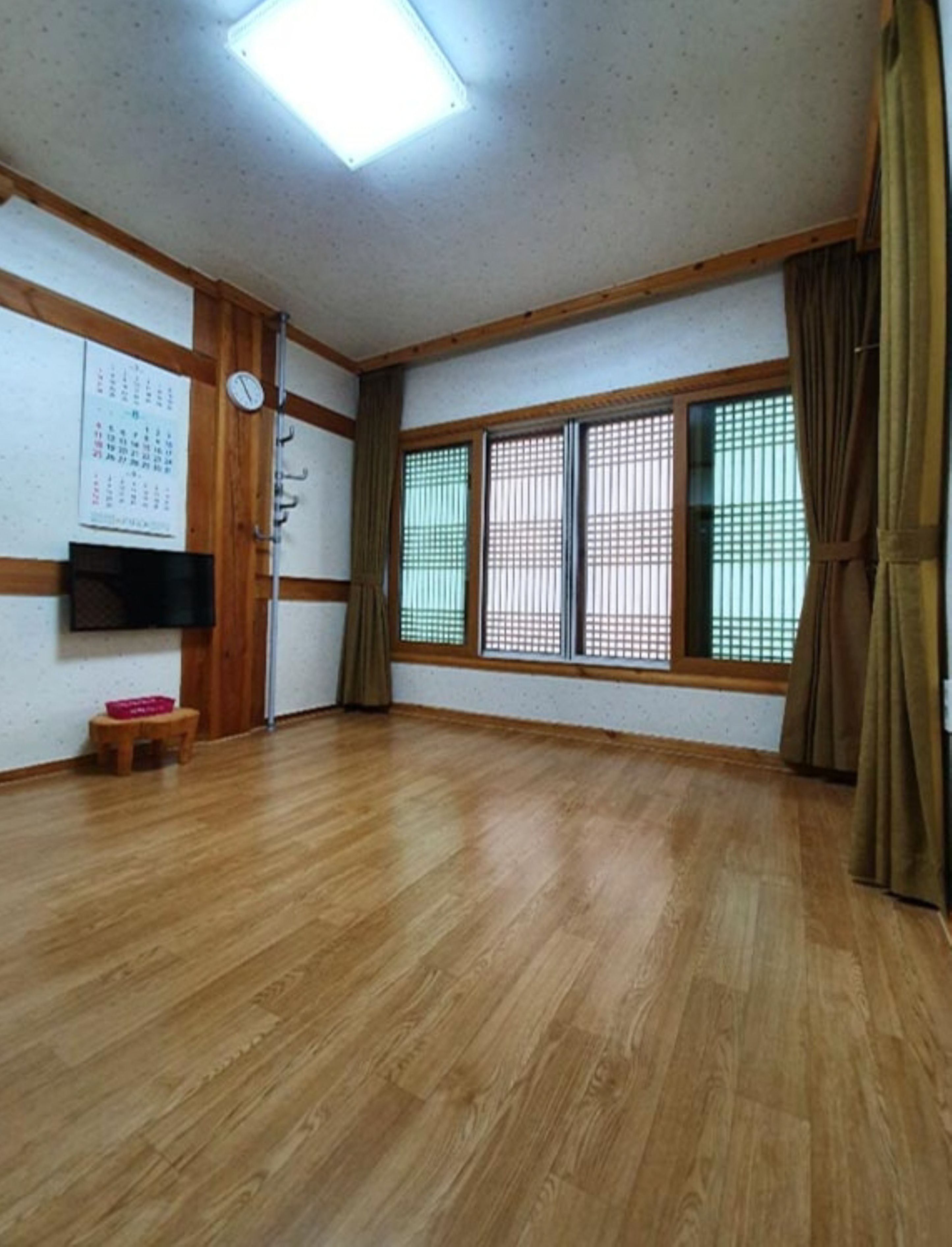 A room with wooden flooring, a flat-screen TV mounted on the wall, a calendar, and a small wooden stool.