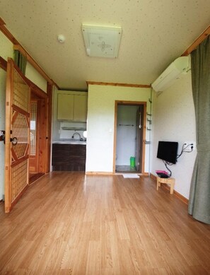 A room with wooden flooring, a ceiling light, a small table with a TV, and a door leading to a kitchen.