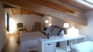 Quarto com teto inclinado, cama, duas cadeiras, mesa de cabeceira com abajures e uma janela.