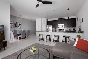 A modern living room with a glass coffee table, a grey sofa, and a dining area with grey chairs.