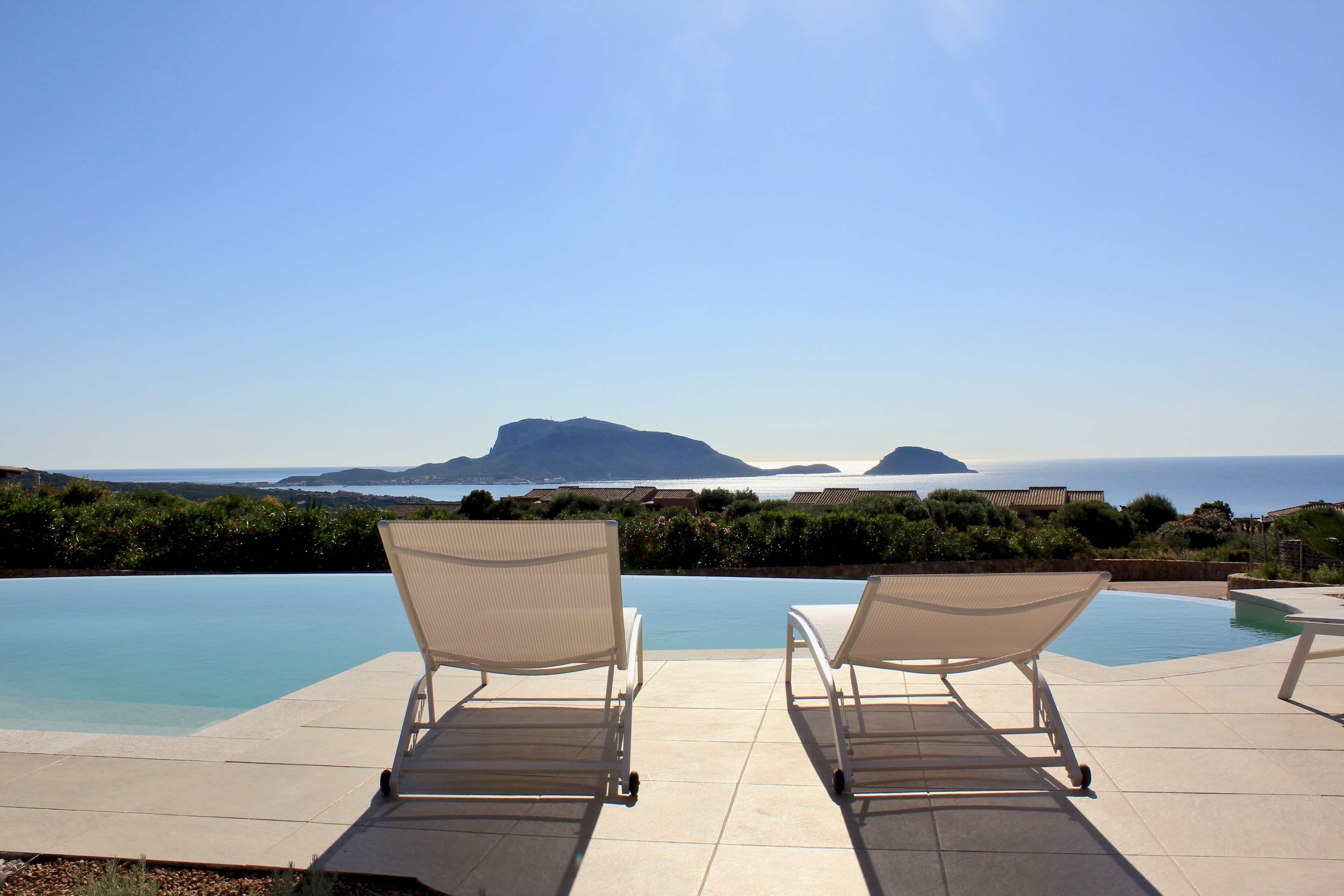 A poolside area with two lounge chairs facing a clear view of the sea and distant mountains.