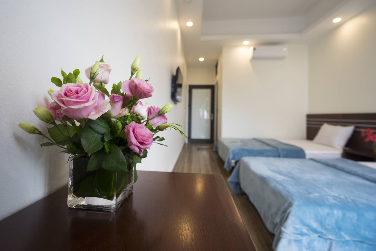 A hotel room with a bed, a bedside table with a vase of pink roses, and a hallway leading to another room.