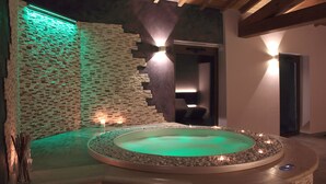 Ein Wellnessbereich mit einem runden Whirlpool, umgeben von einem Kiesstreifen, einer Ziegelwand mit grüner Beleuchtung und einem modernen Sessel im Hintergrund.