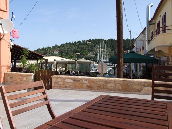 A wooden table and chairs on a patio with a stone wall, overlooking a marina with boats and a hillside in the background.