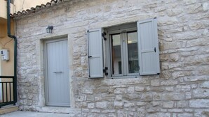 Ein Steinhaus mit zwei weißen Türen und Fensterläden, einer kleinen Treppe zum Eingang sowie einem sichtbaren Stromkasten an der Wand.