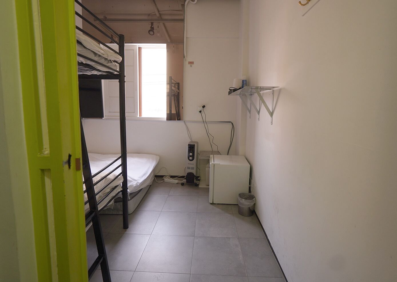 Una habitación pequeña y funcional con literas, un refrigerador compacto, un calentador y una ventana que proporciona luz natural.