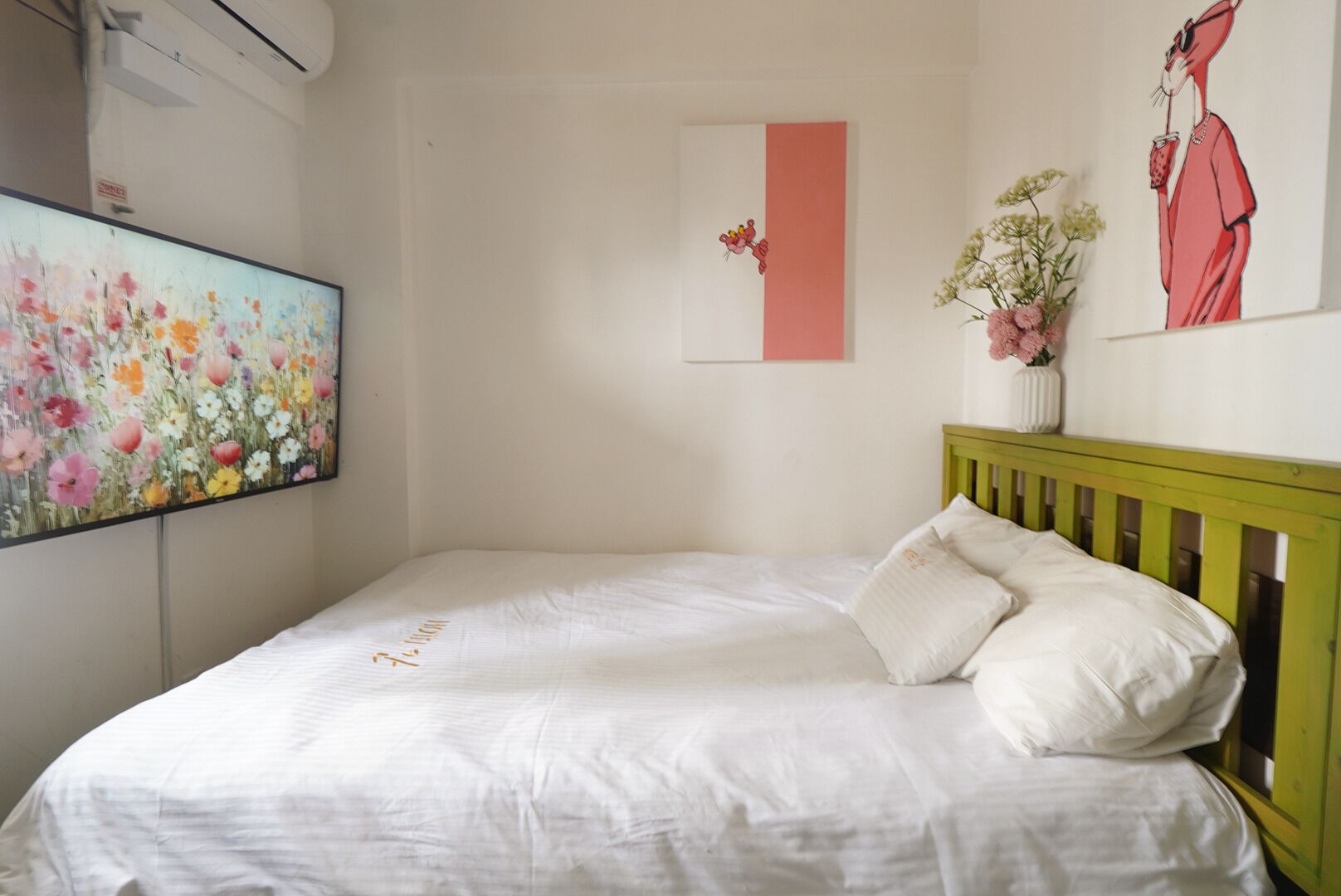 A bedroom with a bed, a wall-mounted air conditioner, a floral painting, a pink accent wall, and a framed illustration.