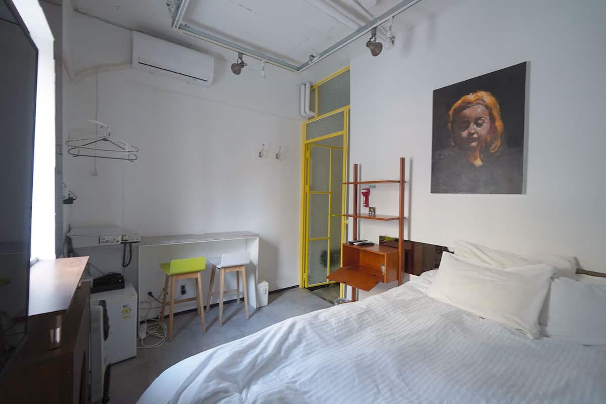 A bedroom with a bed, a desk with two chairs, a mirror, a shelf with a red object, and a painting on the wall.