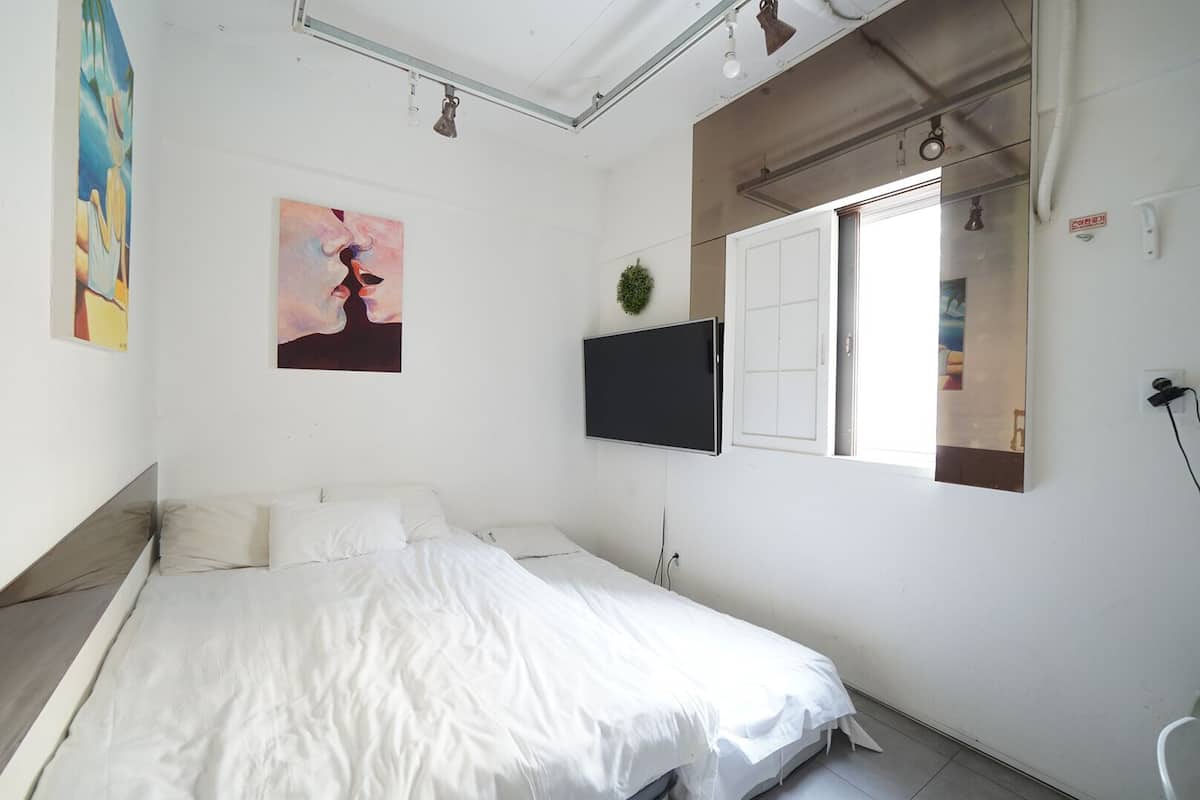 A bedroom with a bed, a TV mounted on the wall, a window, and a painting on the wall.