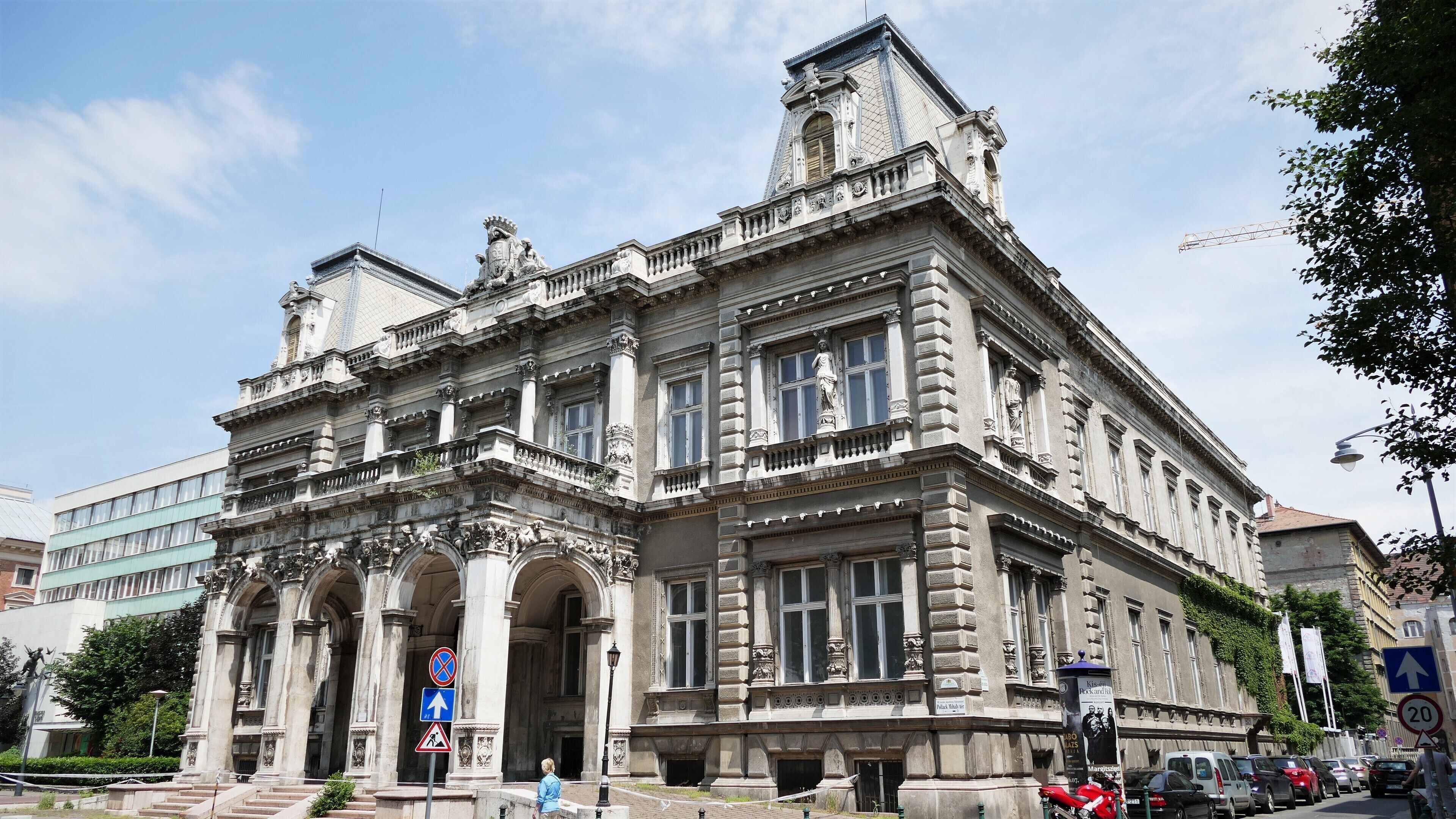 A historic building with ornate architecture, including columns and statues, and a street with cars and a pedestrian.