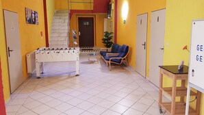 A room with a foosball table, a blue sofa, a small table with a sign, and a door with an exit sign. - Hostel jo-si (Kamenz)