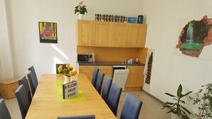 A dining area with a wooden table and blue chairs, a kitchen with wooden cabinets, and a wall with a mural. - Hostel jo-si (Kamenz)