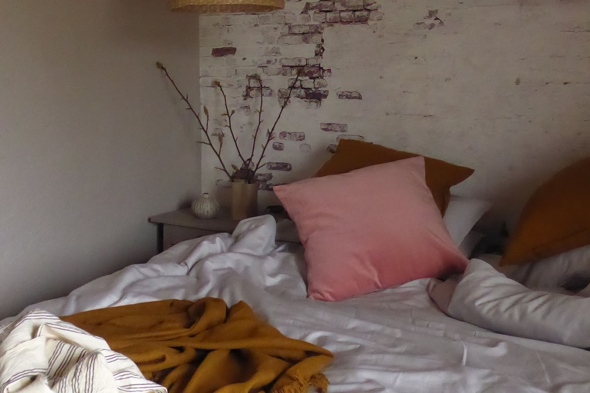 A room with a bed, a hanging lamp, a brick wall, and a small table with a vase.