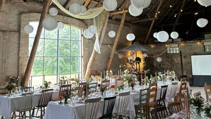 Un espacio rústico con techos altos, vigas de madera a la vista y luces colgantes. Las mesas están dispuestas con manteles blancos y centros de mesa florales.