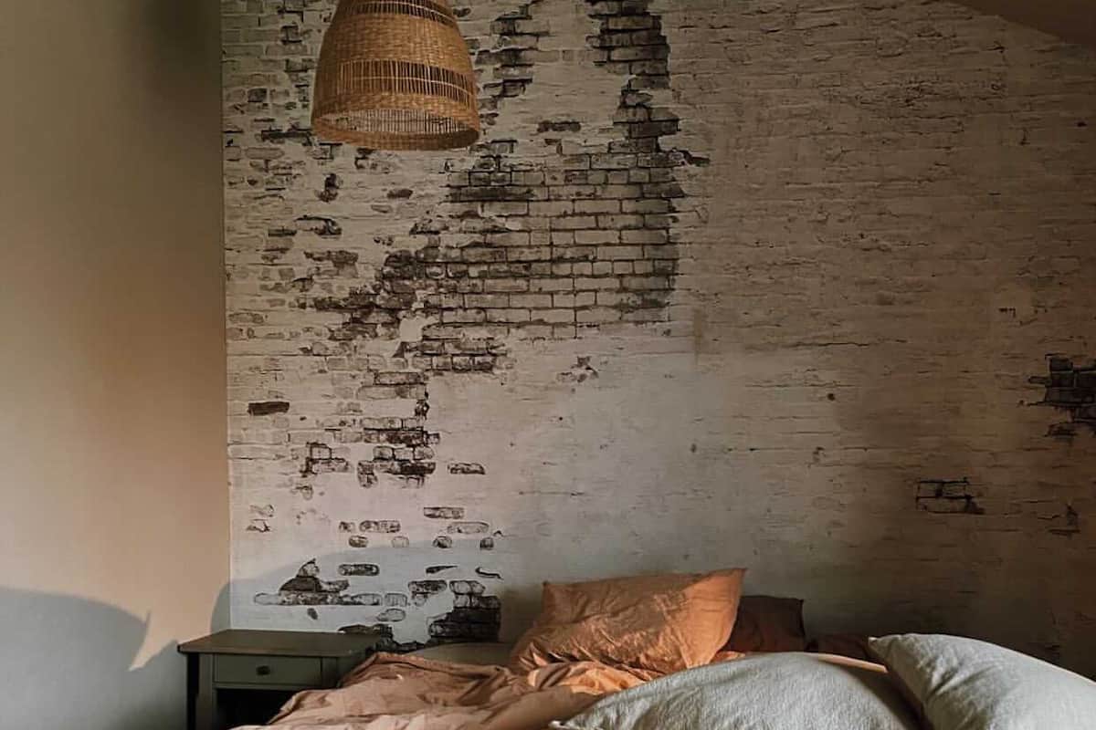 A bedroom with a brick wall, a hanging light fixture, a bed with multiple pillows, and a bedside table with a candle.