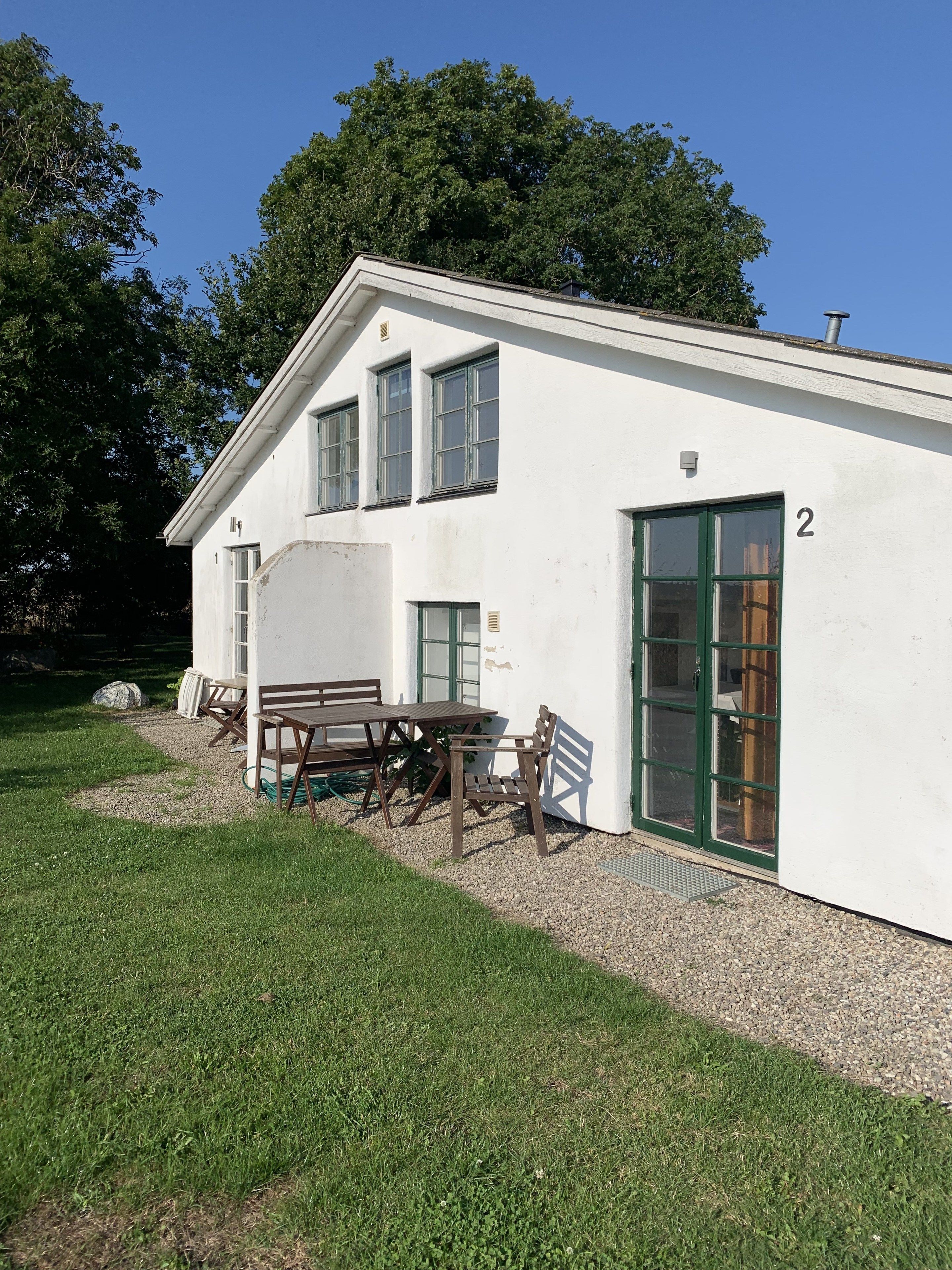 A white building with a number '2' on the side, a gravel driveway, and wooden furniture outside.