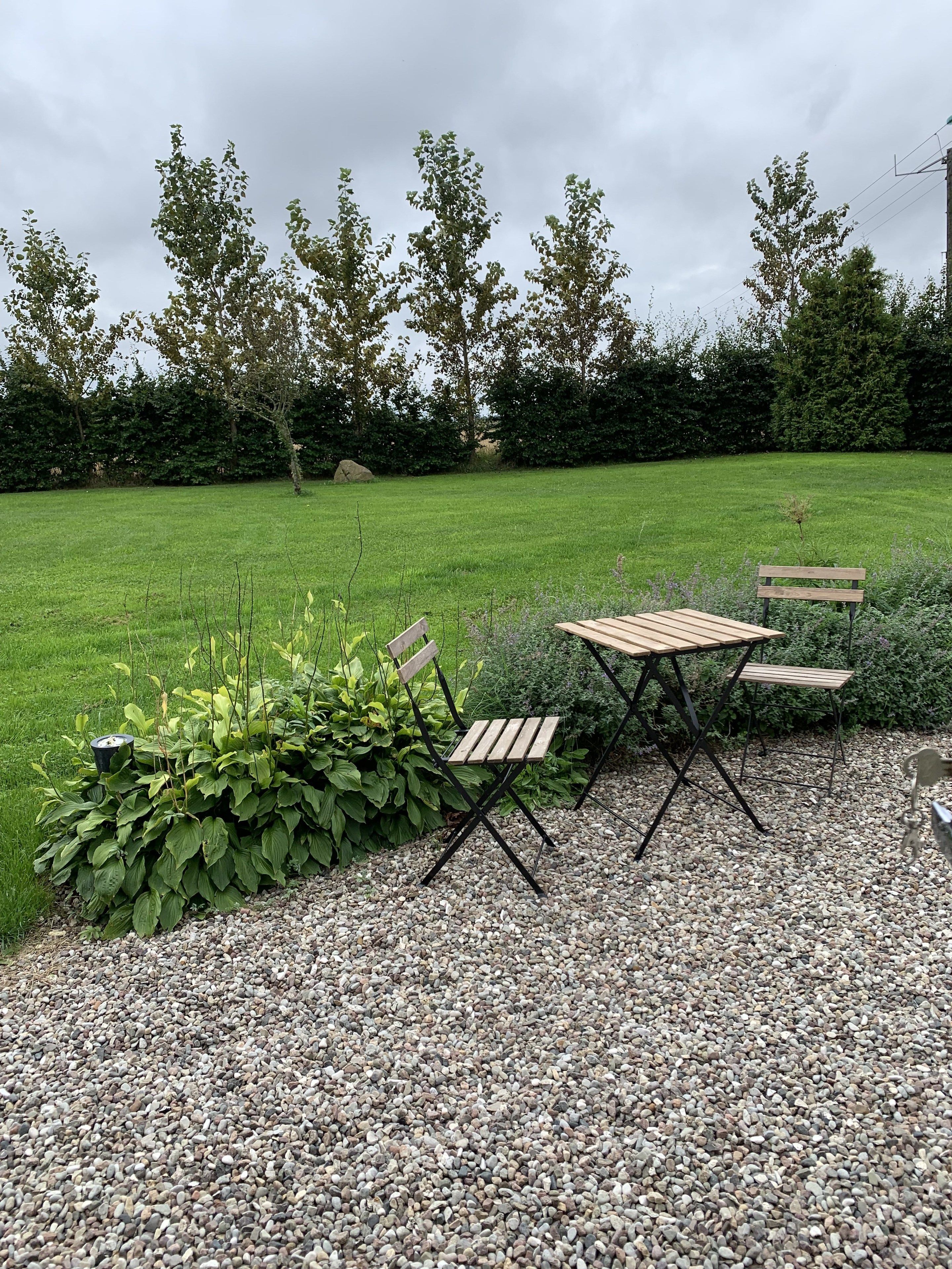 An outdoor seating area with a table and chairs, surrounded by a gravel path and greenery.