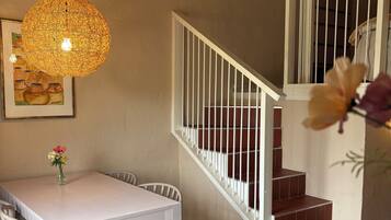 Un comedor con una mesa blanca, sillas, un banco, una lámpara colgante y una escalera con barandillas blancas.