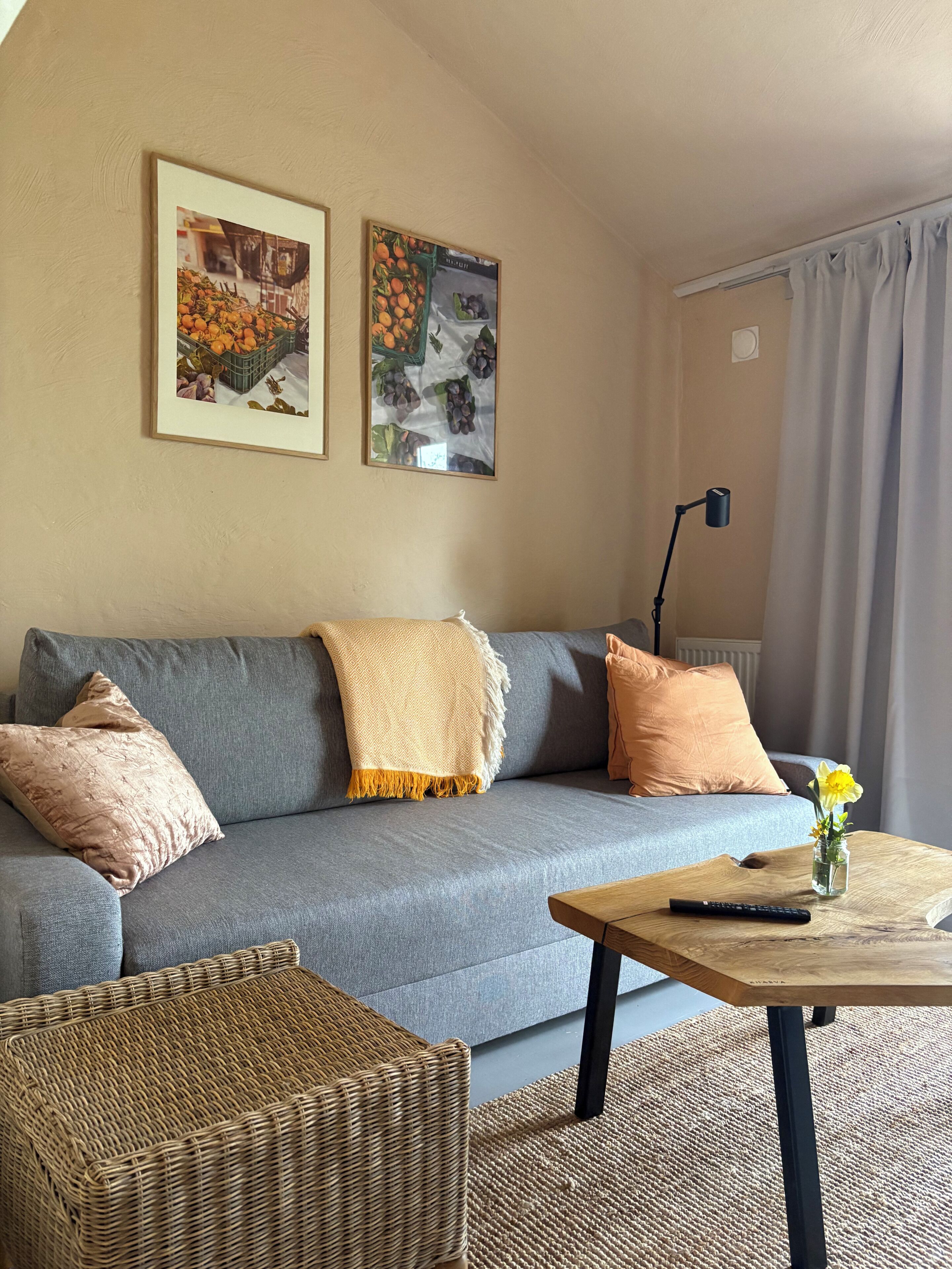 Un salón con un sofá gris, dos cojines, una mesa de centro de madera con un control remoto, un jarrón con flores y dos cuadros en la pared.
