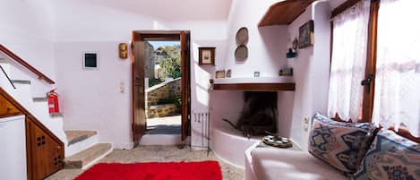 Uma sala de estar acolhedora com tapete vermelho, sofá branco com almofadas estampadas, lareira e vista para uma parede de pedra e vegetação lá fora.
