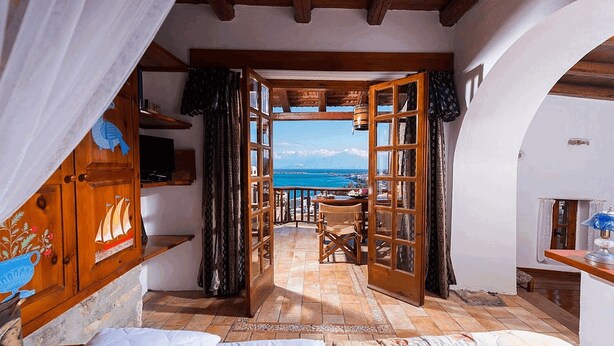 Een kamer met een houten plafond, een bed, een klein tafeltje en uitzicht op zee door een open deur.