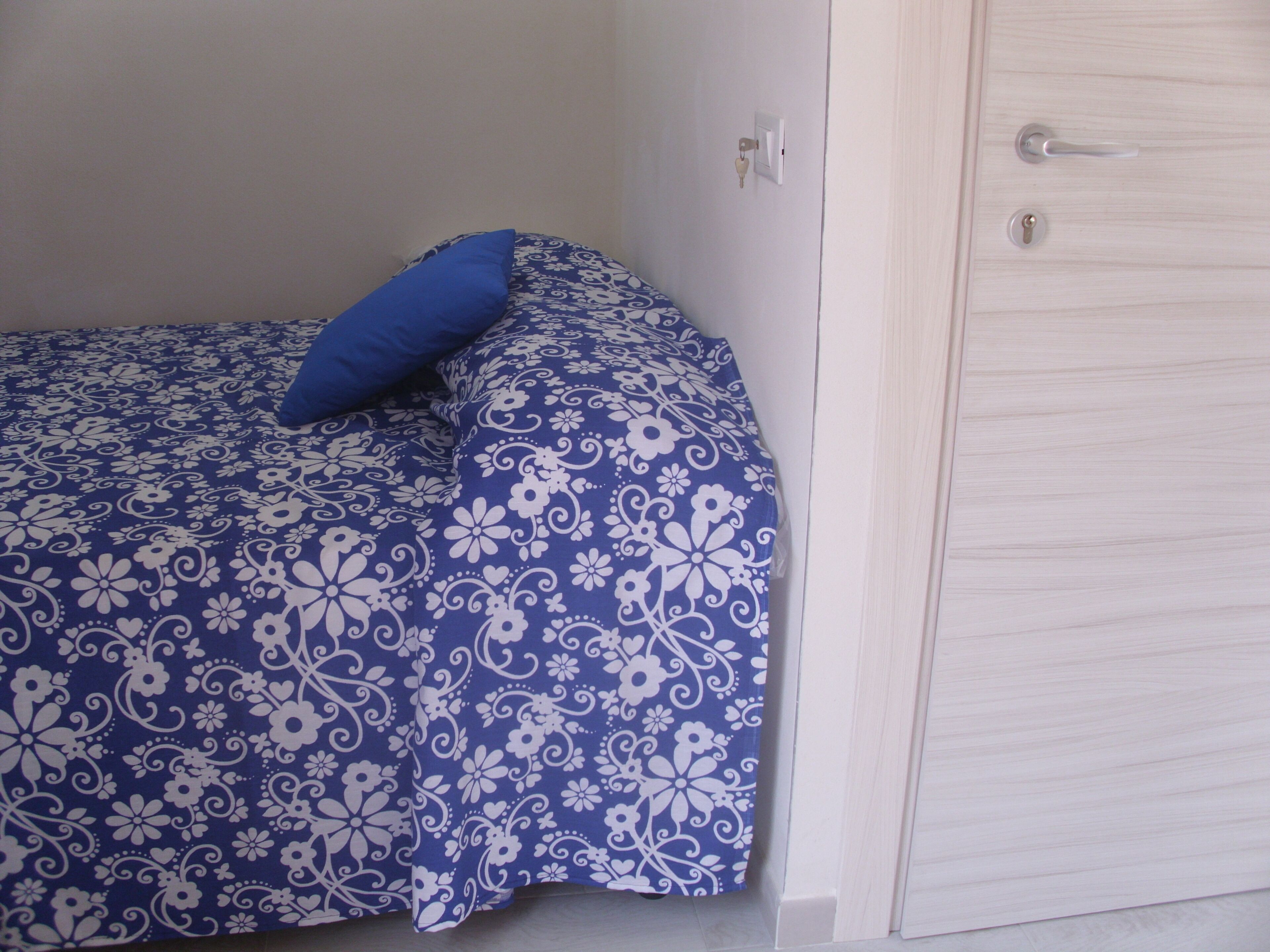 A bedroom with a bed covered in a floral-patterned bedspread, a single blue pillow, and a white wardrobe with a door handle and lock.