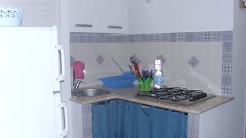 Une cuisine équipée d’un évier, d’un robinet et d’une cuisinière à gaz. Une serviette bleue est accrochée au mur, ainsi qu’une bouteille d’eau et divers ustensiles de cuisine.