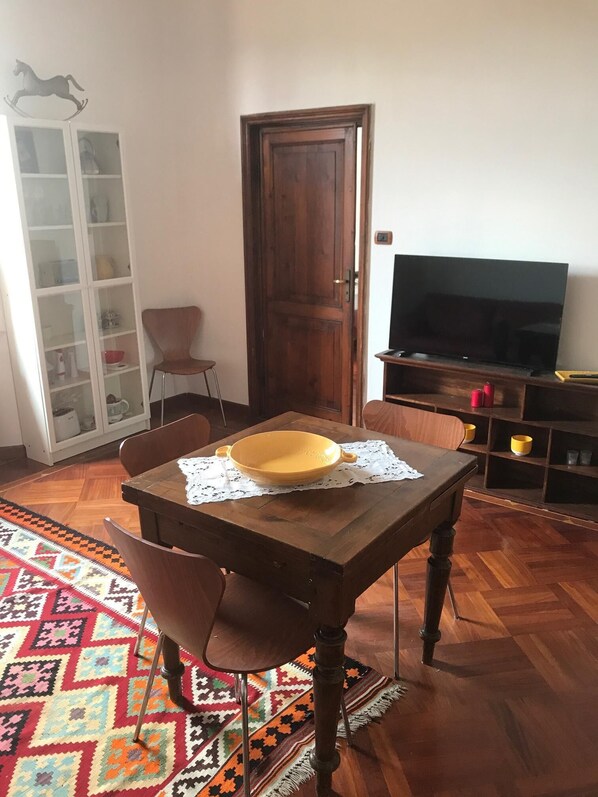 A dining area with a wooden table, chairs, a patterned rug, a cabinet with glass doors, a closed wooden door, and a television on a wooden stand.