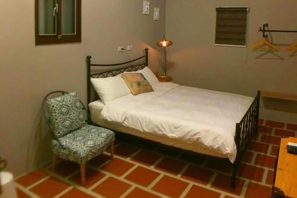 A bedroom with a bed, a chair, a hanging light fixture, and a window with a shutter.