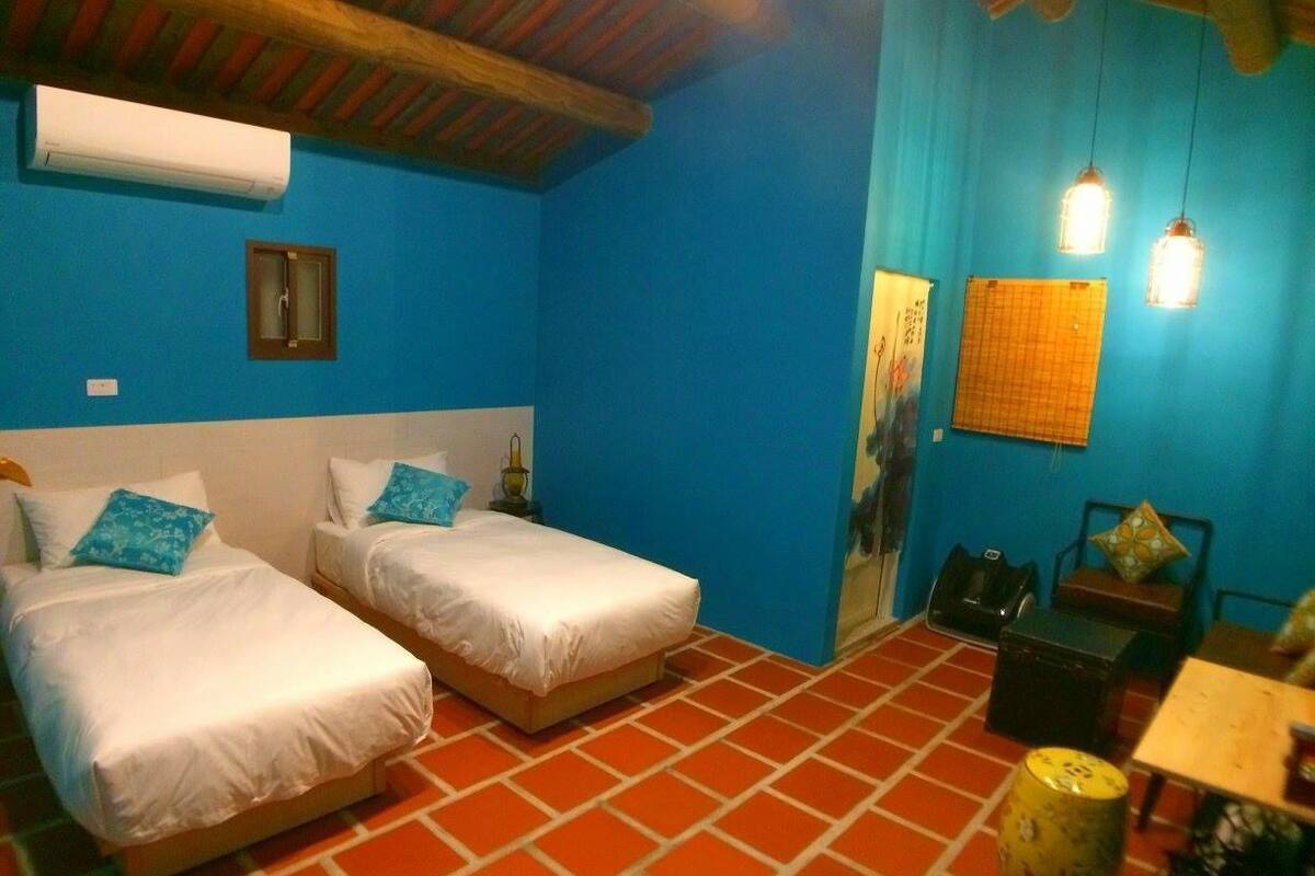 A room with two beds, a table, and a chair. There is a wall-mounted air conditioner and a small window.