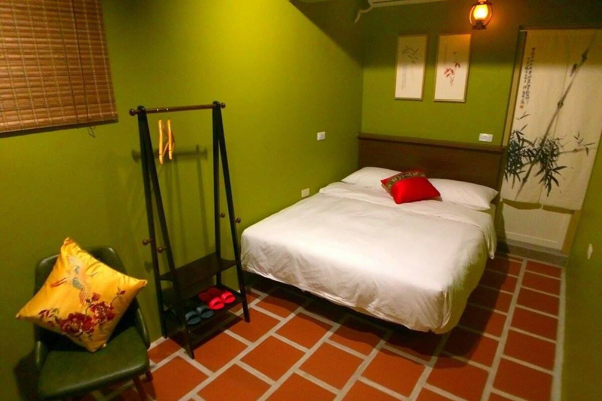 A single bed with a white mattress and red pillow, a green chair with a yellow cushion, a black metal ladder, and a red and white patterned rug.
